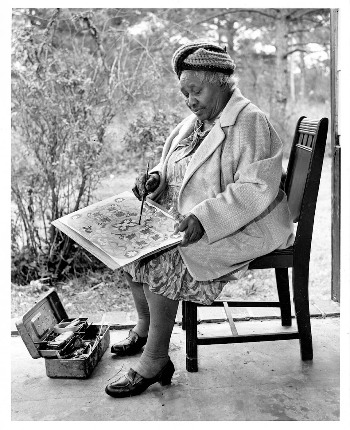 Old black and white photograph of Minnie Evans sitting on chair while drawing.