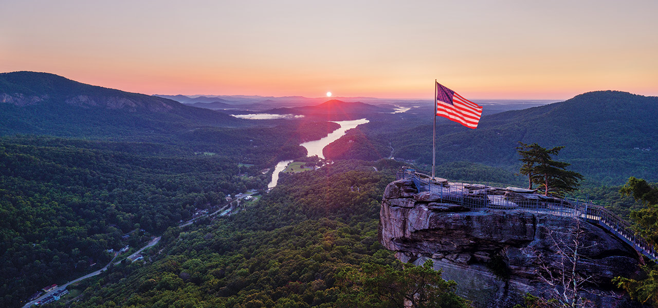 An American flag on top of Chimney Rock, billowing above Lake Lure and Hickory Nut Gorge.